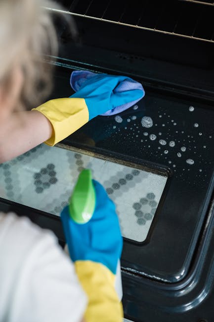 Close-up of a person cleaning the interior of an oven with a blue and yellow rubber glove, holding a green spray bottle in one hand and using a blue cloth to wipe the oven's glass door. The oven has a
