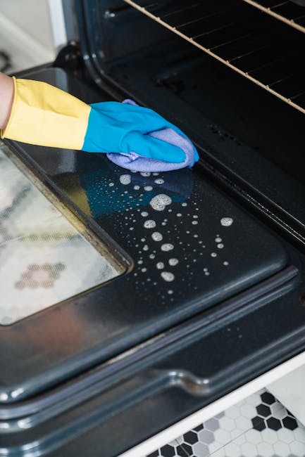 Close-up of a person cleaning the interior of an oven with a blue and yellow rubber glove, holding a green spray bottle in one hand and using a blue cloth to wipe the oven's glass door. The oven has a